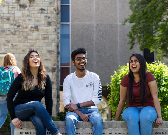 three students sit on a bench outside on 威尼斯人娱乐城 campus. They are laughing.