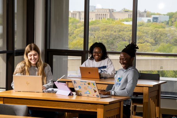 three students sit at tables with open laptops in the library.