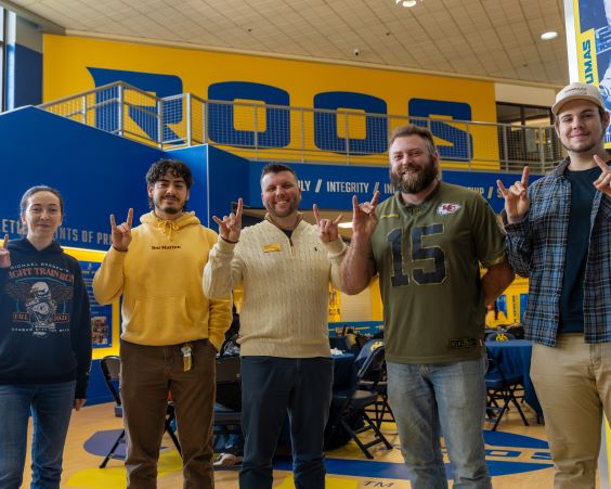 Two students in military blues stand while a student wearing camo scrubs sits in the dental chair in one of the 牙科学院 patient rooms. They are smiling.