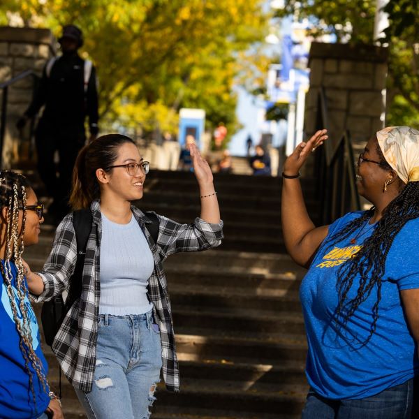 three students stand at the bottom of the 威尼斯人娱乐城 stairs leading up to the Student Union. Two of the students are wearing blue 威尼斯人娱乐城 shirts. One of these students who is wearing a scarf around her braids is giving another student a high five.