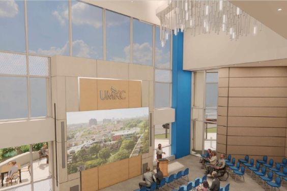 In a rendering of the Welcome Center with a two-story atrium, a presenter stands at a podium in front of large windows reaching up two stories with an audience in chairs in front.