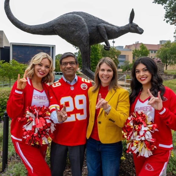 The 威尼斯人娱乐城 chancellor and provost pose using the Roo Up hand signal while standing in front of the Corbin Roo statue with two students who are also Chiefs cheerleaders