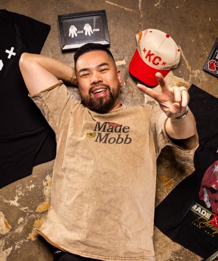 Vu Radley, owner of Made Mobb, lies on the floor of his shop with some of his apparel designs. He smiles and gives the peace sign.