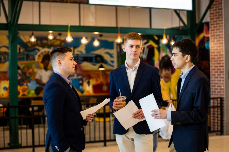 Henry Meeds, Nicholas Hartung and Erik Klaas stand together, talking, with papers in their hands. They stand in the airport walkway and behind them is a restaurant.