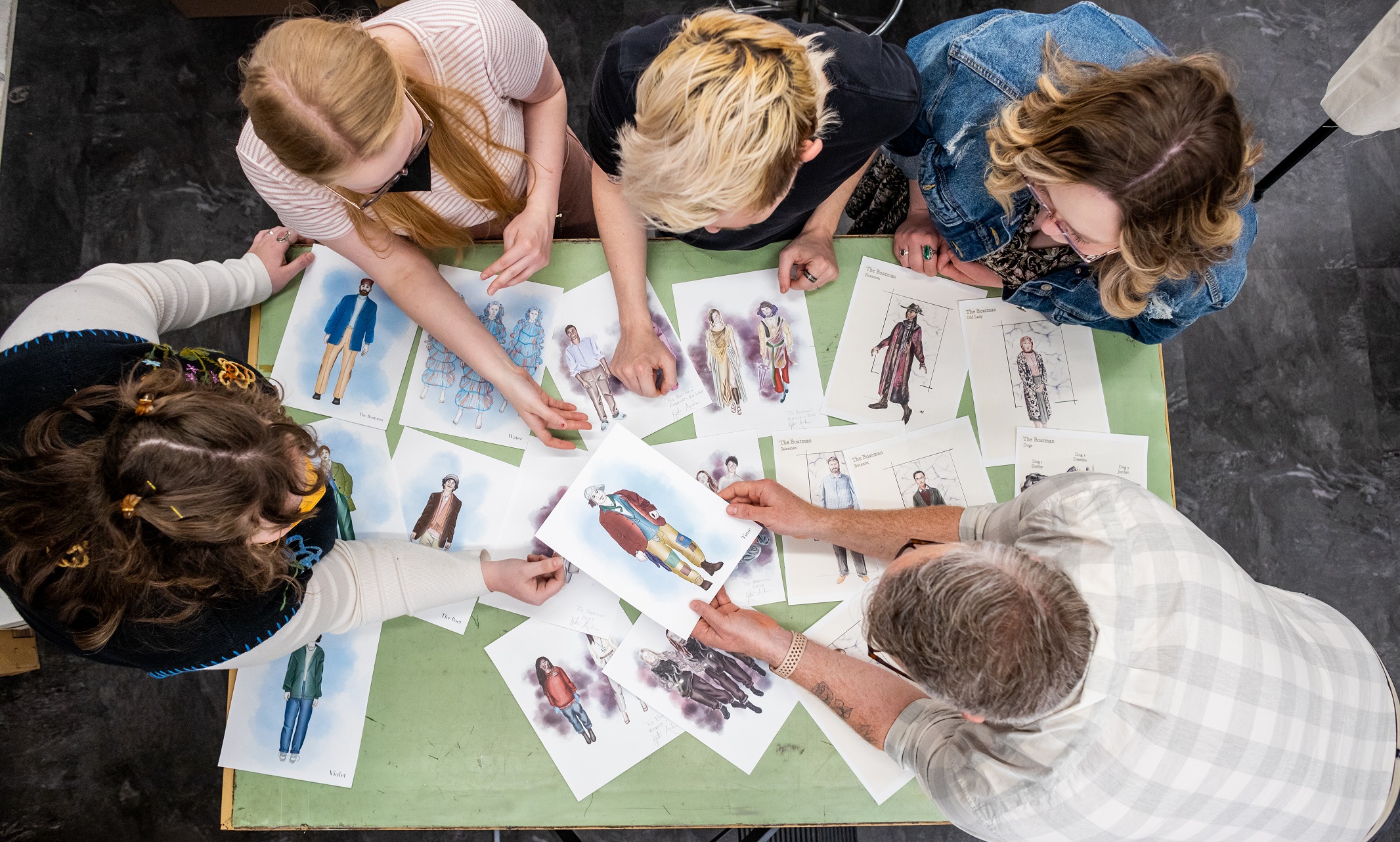 Four students and a professor lean over a light green table. The table is covered with letter paper-sized costume illustrations.