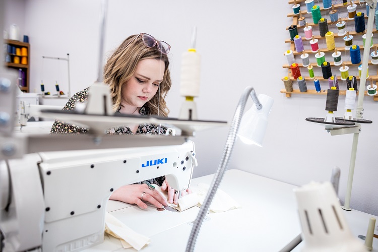 A student sits a sewing machine and works. On the wall beside her is a colorful assortment of thread.