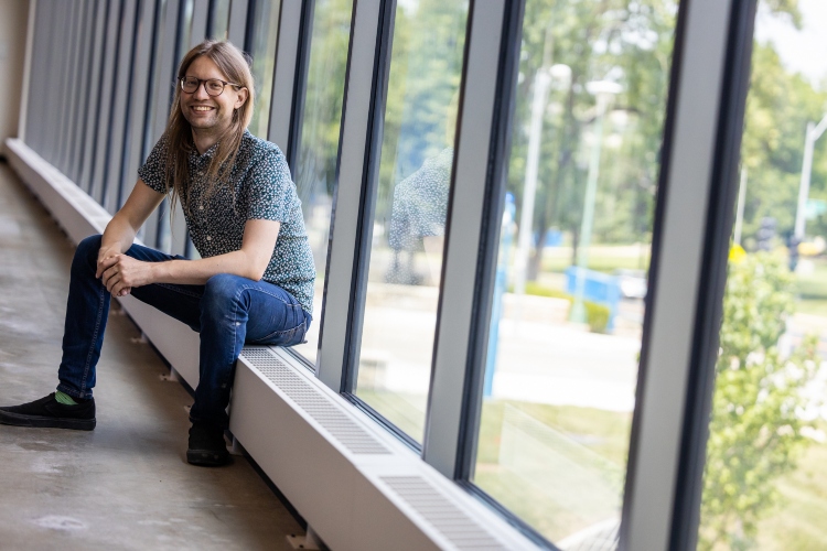 Iain Blair sits in front of a wall of windows inside the Plaster Center with his elbows on his knees and hands clasped in front of him smiling with green space outside the window behind him.