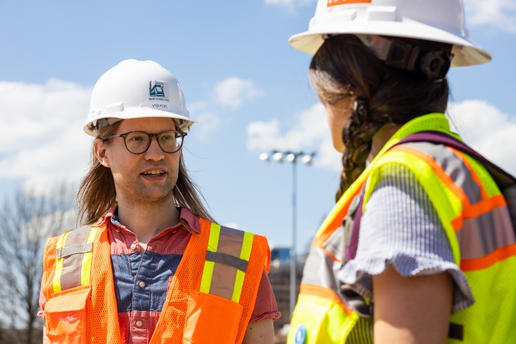 Iain Blair stands in an orange vest and hard hat speaking to a woman with her back to the camera. She is dressed in a yellow construction vest and wearing a hard hat with a braid over her shoulder.