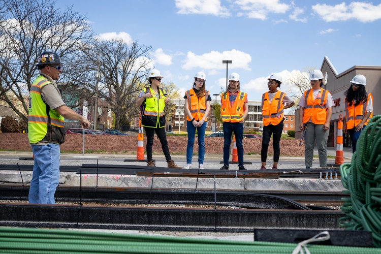A construction worker in a yellow vests stands in a hole filled with lines for the KC streetcar, looking up and speaking to a row of students on the street in orange construction vests and hard hats.