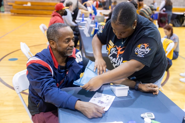 A person helps ano的r person with a blood pressure cuff at a health event inside a gymnasium filled with people