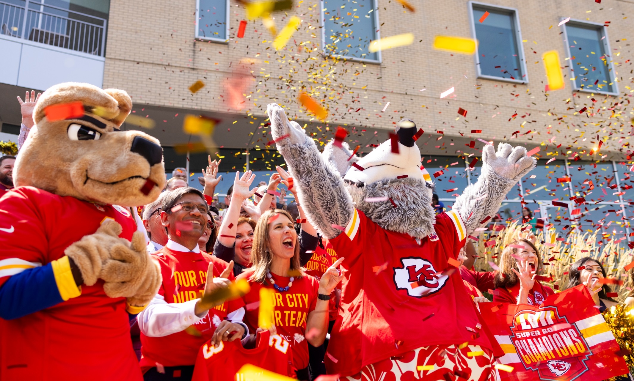 A large group of people, KC Wolf and KC Roo stand in front of the Student Union and cheer enthusiastically as red and gold confetti floats down onto them.