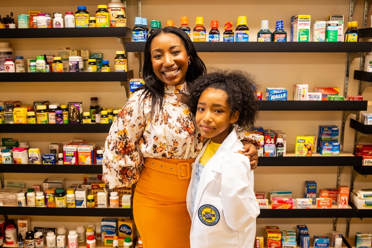 Leresa Cozart stands inside the 威尼斯人娱乐城 Pharmacy mock pharmacy with her arm around her daughter, Mariah, who is wearing her mom's white coat.