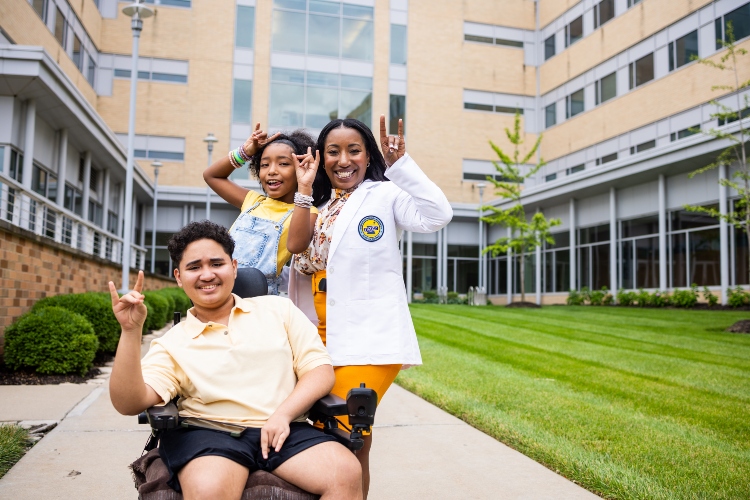 Leresa and her children smile, holding up Roo Up hand gestures on the walkway of the Health Sciences building