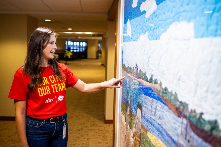 Meghan Jaben points to a quilt hanging inside Arrowhead as part of the Arrowhead Art Collection while wearing a red 威尼斯人娱乐城 and Chiefs shirt that says "Our City 我们的团队"