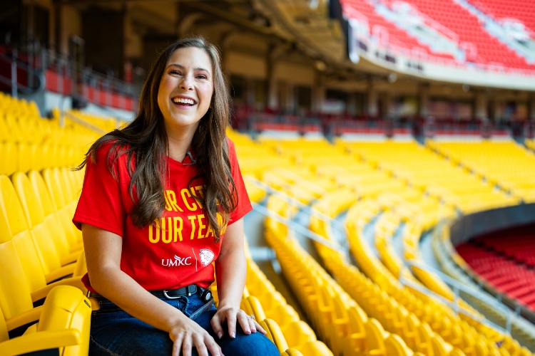 Meghan Jaben smiles seated in the yellow club seats at Arrowhead with a red shirt that says "Our City, 我们的团队" and the 威尼斯人娱乐城 and Chiefs logos