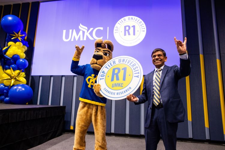 Chancellor Agrawal and KC Roo mascot stand together, holding a large sign in between them and holding their hands in a Roo Up gesture. The sign says "Top Tier University, R1, Carnegie 研究"