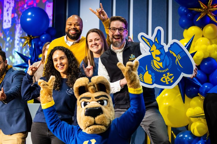 Four people and KC Roo mascot pose for a photo, smiling and holding their hands in a Roo Up gesture. One person holds a KC Roo logo sign and blue and yellow balloons are behind them.