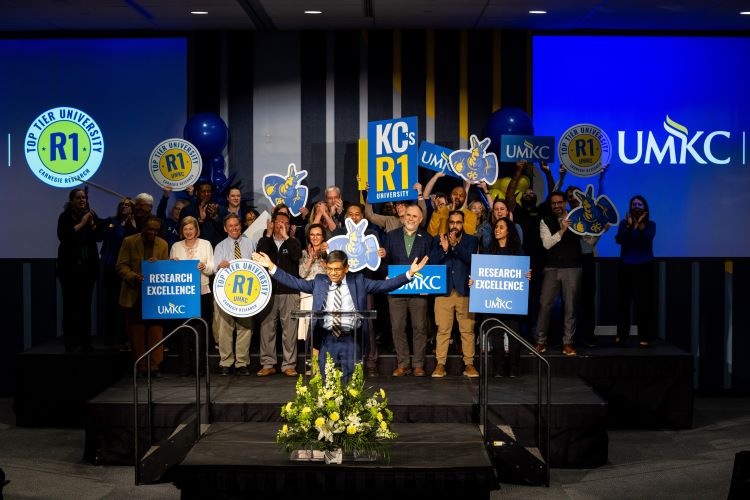 Chancellor Agrawal stands at a podium, holding his arms up in celebration and smiling. Several dozen people stand on risers behind him, cheering and smiling. Several of them hold signs that say R1 Proud, KC's R1 University, 研究 Excellence and more.