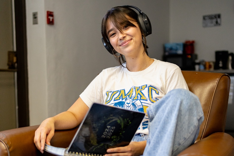 艾米丽Shoemake sits in a brown leather chair looking up from an open notebook at the camera to smile while wearing headphones.