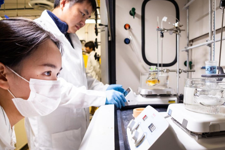 A student and a professor wearing white lab coats conduct an experiment under a flume hood.