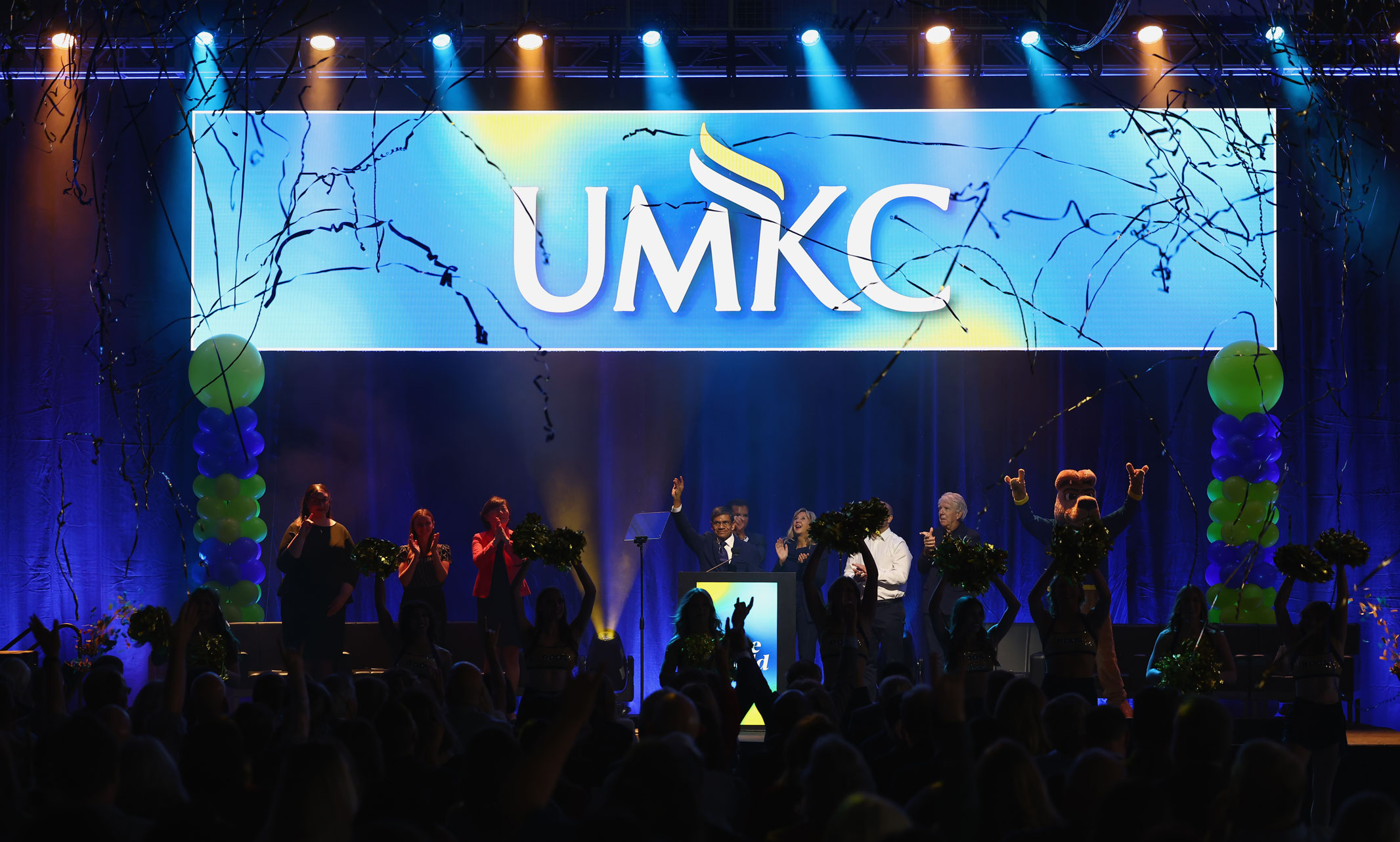 A group of people stand on stage with blue and gold streamers and confetti falling from the ceiling.