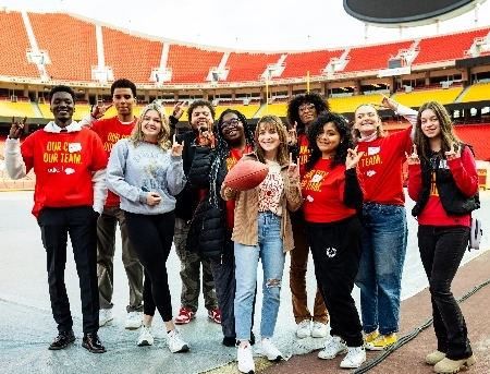 Three people walk through football practice complex. Two are wearing red shirts. One person is wearing jeans and olive green 首领 shirt