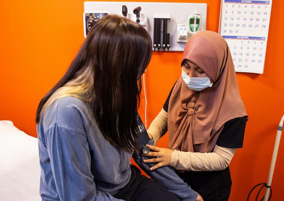 Person standing wearing facemask is holding person sitting on medical office bed's arm as they apply the blood pressure cuff
