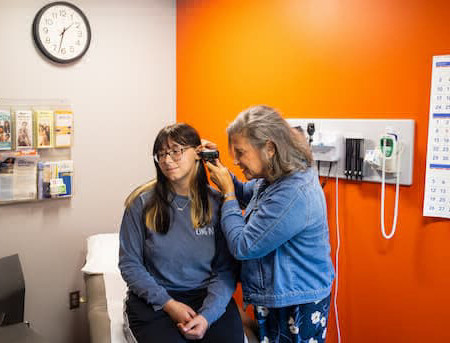 Person on right is squinting and looking into the ear of person in medical office. 