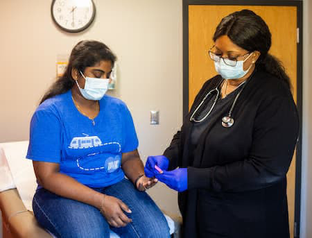 Roo健康 staff member wearing scrubs and mask is pricking the finger of a patient who is also wearing a mask.