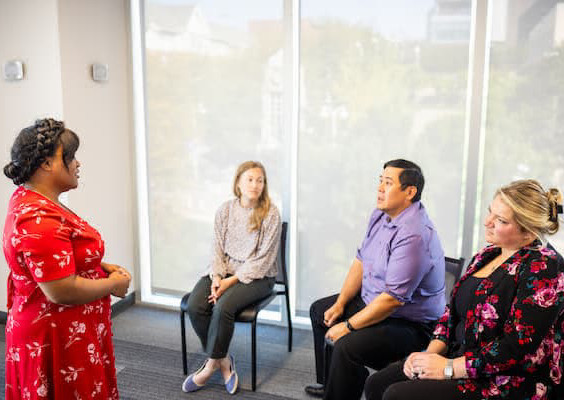 Person in red flower dress with brown hair pulled back is standing and facing three people sitting in chairs..
