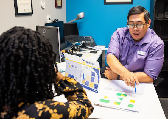 Person wearing glasses and blue button down shirt points with pin at piece of paper. Another person with back to camera sits at same desk with flower shirt.
