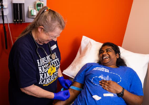 Person laying down on medical office bed is getting arm wrapped by person wearing a 堪萨斯城 shirt and wearing gloves and stethoscope